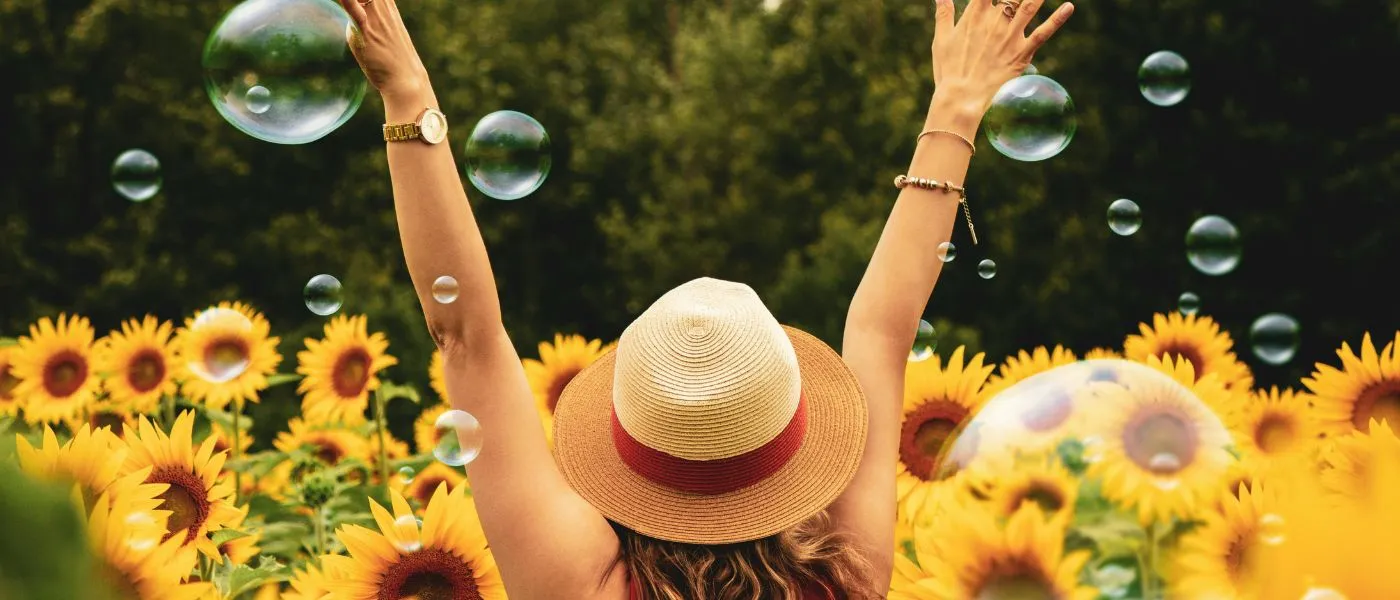 Person mit Sonnenhut hebt in einem Sonnenblumenfeld die Arme.