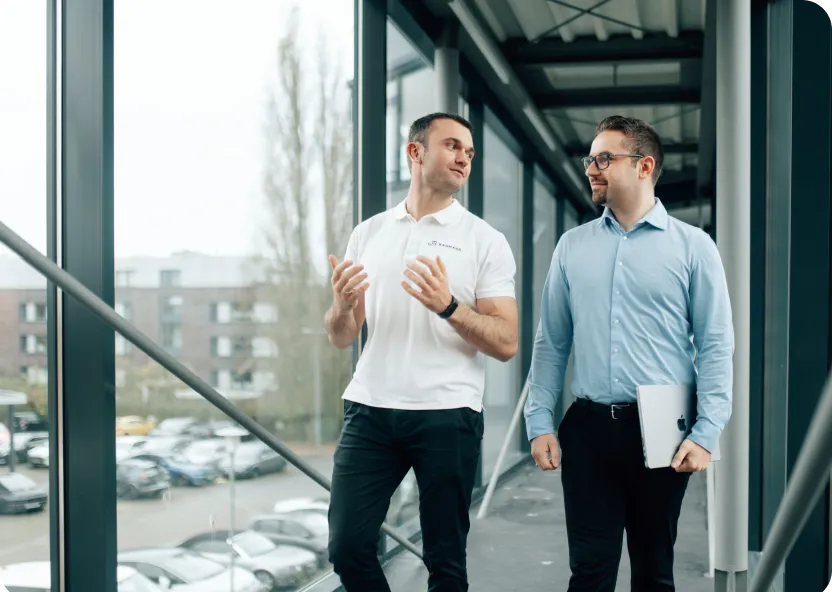 Zwei Männer gehen durch einen verglasten Büroflur und unterhalten sich angeregt. Einer trägt ein weißes „Bahmann“-Poloshirt und gestikuliert beim Sprechen, der andere hält ein MacBook und hört aufmerksam zu.