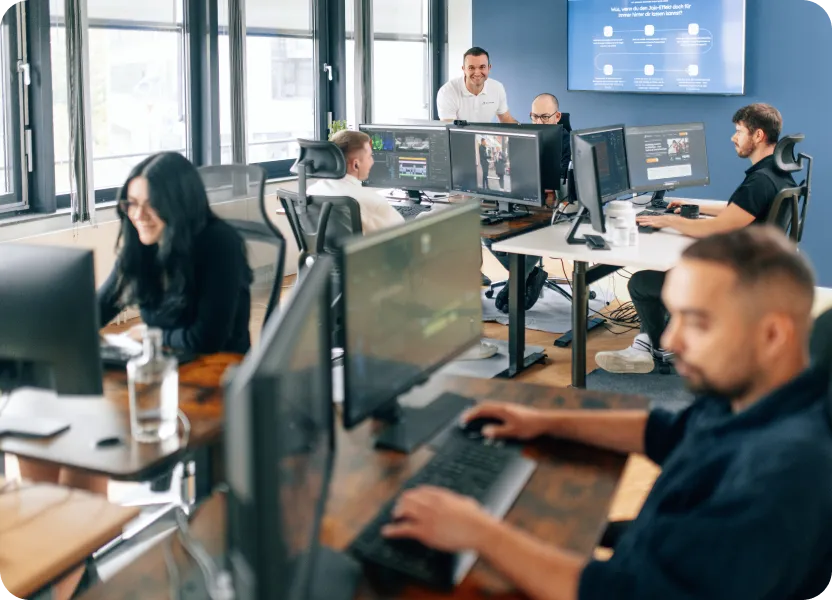 Ein kreatives Team arbeitet konzentriert in einem modernen Büro mit mehreren Monitoren und großen Fenstern. Im Hintergrund steht ein lächelnder Kollege im weißen „Bahmann“-Shirt, während auf einem großen Bildschirm an der Wand eine Präsentation angezeigt wird.