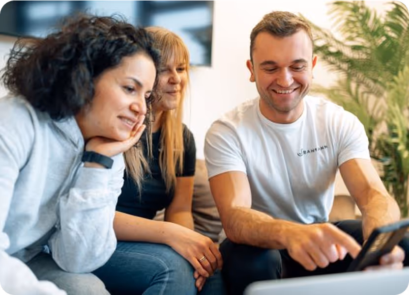 Drei Personen sitzen entspannt auf einem Sofa in einem hellen Raum und schauen gemeinsam auf ein Smartphone, das von dem Mann rechts in der Hand gehalten wird. Alle wirken gut gelaunt; der Mann trägt ein weißes „Bahmann“-T-Shirt, während die beiden Frauen interessiert lächeln.