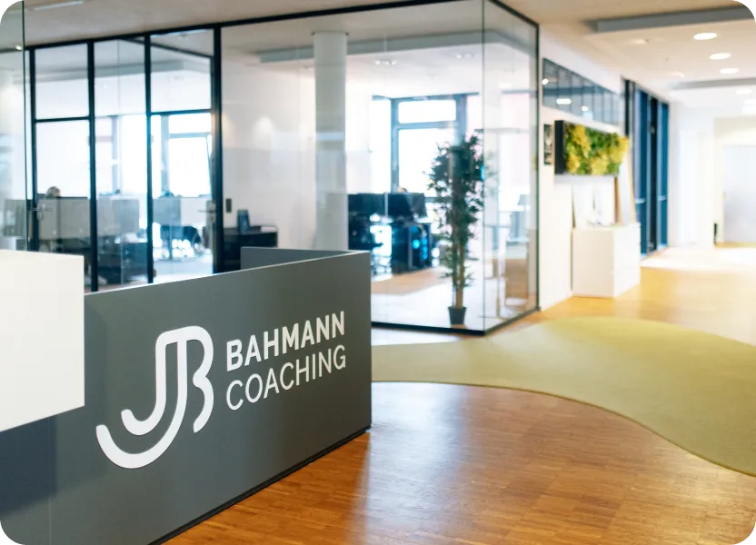 Helles, modernes Büro mit Glaswänden und einer Empfangstheke, auf der das Logo „JB Bahmann Coaching“ zu sehen ist. Im Hintergrund befinden sich Arbeitsplätze, eine bepflanzte Wanddekoration und ein einladender Flurbereich mit Holz- und Teppichboden.