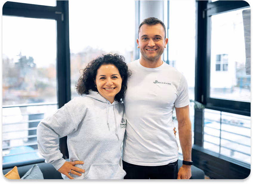 Ein Mann und eine Frau stehen Arm in Arm in einem modernen Büro mit großen Fenstern und lächeln in die Kamera. Beide tragen Kleidung mit dem „Bahmann“-Logo – er ein weißes T-Shirt, sie einen grauen Hoodie.