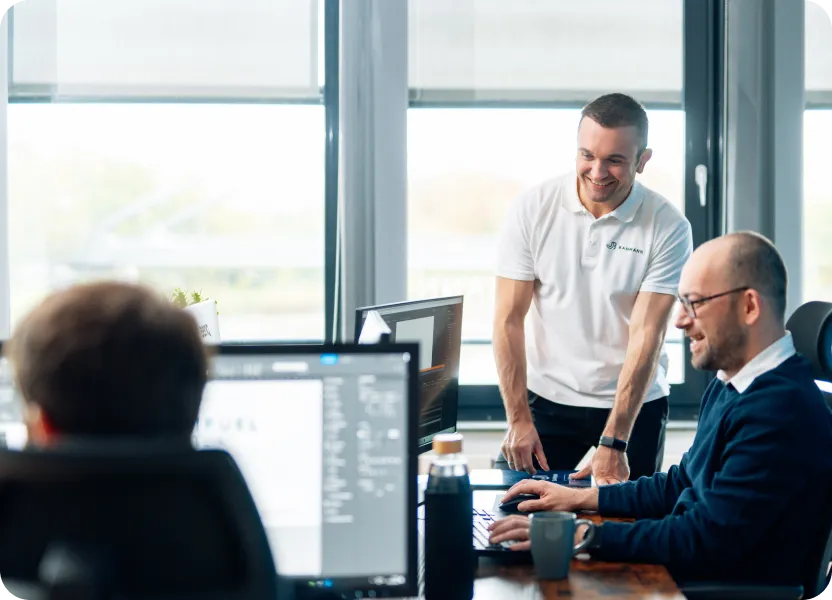 In einem modernen Büro arbeiten mehrere Männer konzentriert an ihren Monitoren, während ein Kollege im weißen „Bahmann“-Poloshirt lächelnd bei einem Arbeitsplatz steht und ins Gespräch vertieft ist. Die Szene wirkt freundlich und produktiv, mit viel Tageslicht im Raum.