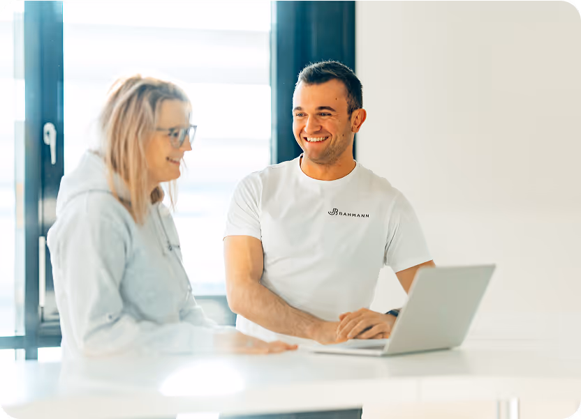 Zwei Kolleg:innen stehen an einem weißen Tisch in einem hellen, modernen Büro und unterhalten sich freundlich. Der Mann im weißen „Bahmann“-T-Shirt arbeitet an einem Laptop, während die Frau im grauen Hoodie lächelt und zuhört.