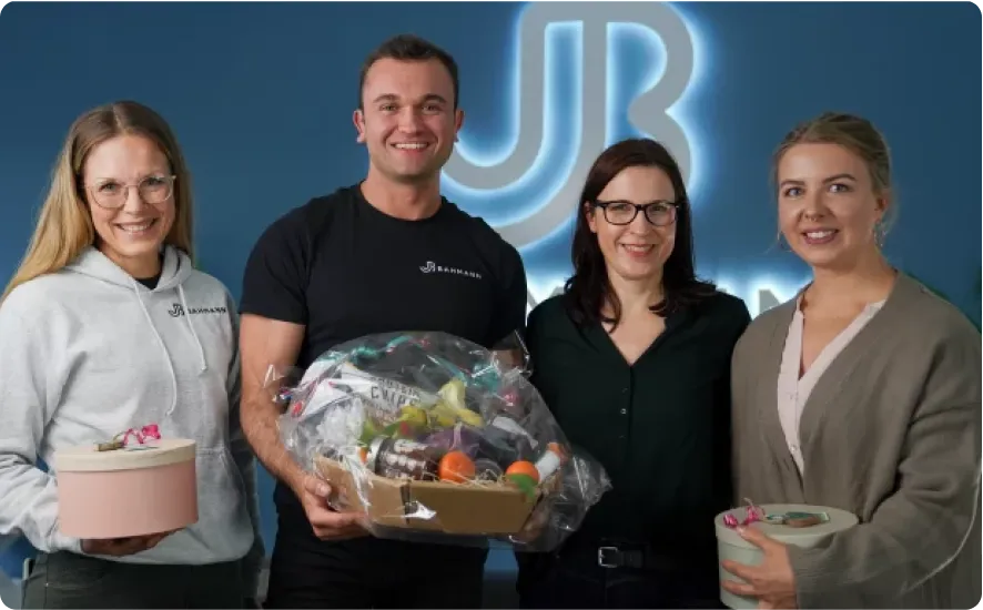 Jan Bahmann steht mit drei Frauen in einem Büro vor einem leuchtenden JB-Logo. Sie halten Geschenke und präsentieren sich lächelnd für das Foto, das ein erfolgreiches und freundliches Arbeitsumfeld symbolisiert.